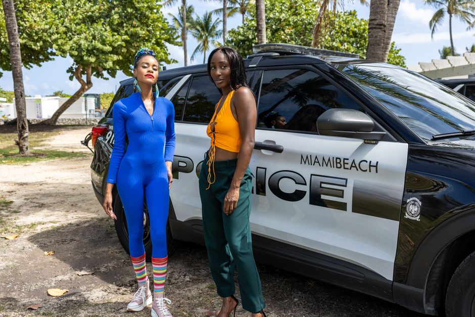 Two stylish women stand beside a Miami Beach police car in a sunny outdoor setting.