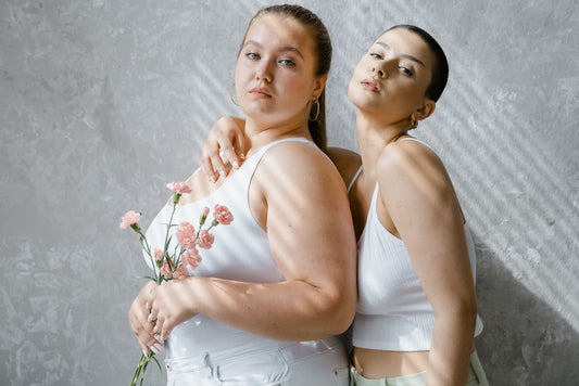 Two confident women stand together holding pink flowers in a modern indoor setting.